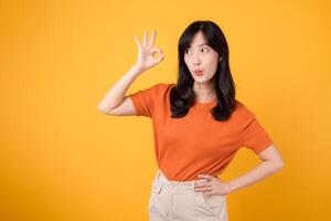 Radiating positivity, young Asian woman 30s showcases okay sign in orange shirt on vibrant yellow background. Hands gesture concept photo