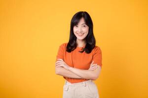 Confident young Asian woman in her 30s, wearing an orange shirt, showcases crossed arm sign gesture on yellow background. Hands gesture concept. photo
