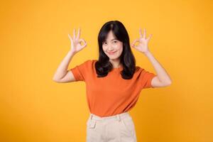 A confident young Asian woman in her 30s, dressed in an orange shirt, displays the okay sign gesture on a bright yellow background. Hands gesture concept. photo