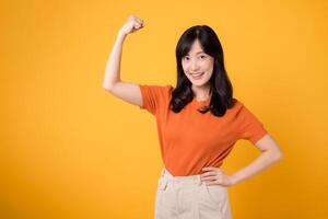 Confident young Asian woman in her 30s raises a fist up hand sign gesture, wearing an orange shirt on yellow background. Empowerment and celebration concept. Empowerment and feminism concept. photo