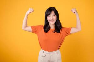 Confident young Asian woman in her 30s celebrates with a fist up hand sign gesture, wearing an orange shirt on yellow background. Empowerment and feminism concept. photo