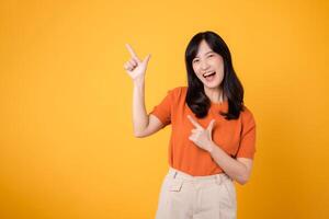 Embody joy with a young Asian woman in her 30s, donning an orange shirt, pointing to free copy space on yellow background. recommending product concept. photo