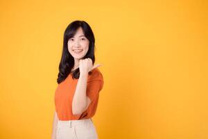 Experience happiness with a young Asian woman in her 30s, donning an orange shirt, pointing her thumb finger to free copy space on yellow background. product recommending concept. photo