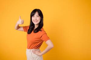 Celebrate happiness with a young Asian woman in her 30s, wearing an orange shirt, giving a thumbs up hand gesture on yellow background. product recommending concept. photo