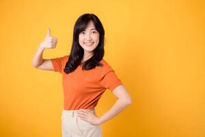 Young asian woman 30s wearing orange shirt showing thumb up hand gesture isolated on yellow background. cheerful happy face female person with hands gesture and product recommending concept. photo