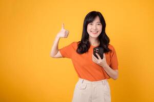 Experience happiness with a young Asian woman in her 30s, donning an orange shirt, showing thumb up gesture while using smartphone on yellow background. photo