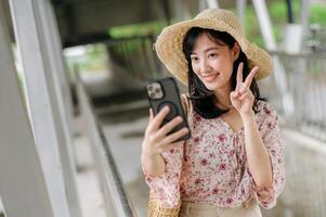 young asian woman traveler with weaving basket selfie with mobile phone and standing on overpass. Journey trip lifestyle, world travel explorer or Asia summer tourism concept. photo