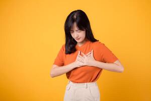 Attentive young Asian woman in her 30s, wearing an orange shirt, holds hands on chest on yellow background. heart attack disease, chest pain health care concept. photo