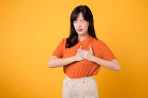 Caring young Asian woman in her 30s, wearing an orange shirt, holds hands on chest on yellow background. heart attack disease, chest pain health care concept. photo