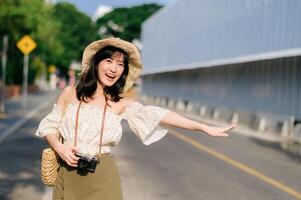 Portrait of asian young woman traveler with weaving hat and basket and a camera waving hand to friend by the street. Journey trip lifestyle, world travel explorer or Asia summer tourism concept. photo