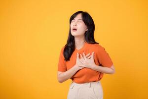 Concerned Asian woman in her 30s, wearing an orange shirt, holds hands on chest on yellow background. heart attack disease, chest pain health care concept. photo