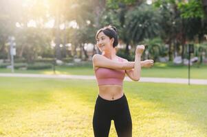 bienestar y sano estilo de vida retrato 30s asiático mujer en rosado ropa de deporte. preparando y extensión cuerpo y brazo músculos antes de un puesta de sol correr en el parque. aptitud fuera de y abrazo equilibrado vida. foto