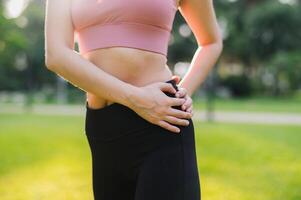 cerca arriba ajuste hembra persona que practica jogging como un asiático mujer en su 30, vistiendo rosado ropa de deporte, calienta abajo y sostiene cadera después un puesta de sol correr en un público parque. abrazo el concepto de bienestar vivo y naturaleza. foto