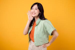 Energetic young Asian woman 30s wearing a green shirt on an orange background, enthusiastically shouting with excitement. Explore the concept of discount shopping promotion with this vibrant image. photo