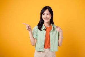 joven asiático mujer 30s vistiendo un verde camisa en un naranja camisa, con confianza señalando dedos a gratis Copiar espacio. explorar el concepto de descuento compras promoción con atractivo imagen. foto