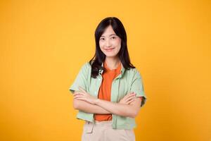 young Asian woman 30s, wearing green shirt on orange shirt with happy face and crossed arm gesture. confident female concept against yellow backdrop. photo