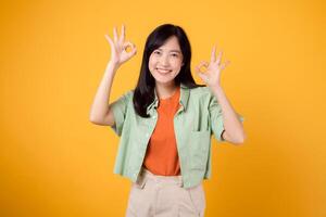 delightful young Asian woman 30s, dressed elegantly in orange shirt and green jumper. Her expressive okay hand gesture and gentle smile shine brightly on yellow background, body language concept. photo