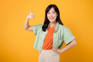 positive feedback of approval with young Asian woman 30s, dressed gracefully in orange shirt and green jumper. Her expressive thumbs up gesture, captured on yellow background, concept of acceptance. photo