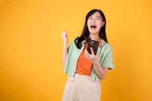 Discover the joy of great news with a cheerful young Asian woman in her 30s, wearing orange shirt and green jumper, using smartphone with fist up gesture on yellow background. Mobile concept. photo