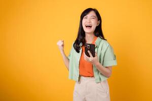 Experience the excitement of great news with a vibrant young Asian woman in her 30s, donning orange shirt and green jumper, using smartphone with fist up gesture on yellow background. Mobile concept. photo