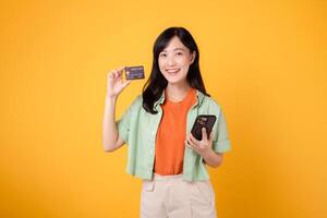 descubrir el alegría de en línea compras con alegre joven asiático mujer en su 30, vistiendo naranja camisa y verde saltador, utilizando teléfono inteligente a espectáculo crédito tarjeta en amarillo estudio antecedentes. móvil concepto. foto
