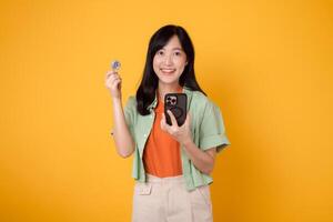 young 30s asian woman happy face dressed in orange shirt and green jumper showing crypto currency coin while holding smartphone isolated on yellow background. Future finance concept. photo