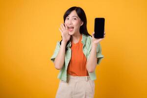 Happy young Asian woman 30s, wearing orange shirt and green jumper, presents smartphone screen with shout mouth on yellow background. New mobile application, new mobile device concept. photo