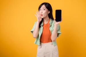 cheerful young Asian woman 30s, donning orange shirt and green jumper, showcasing smartphone screen with a shout mouth on yellow studio background. New mobile device and online shopping concept. photo