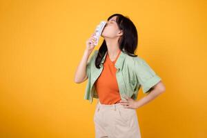 financial money with cheerful young Asian woman 30s, donning orange shirt and green jumper, kissing dollar currency while striking an akimbo gesture on yellow background. Financial money concept. photo