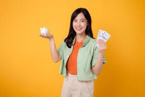 alegre joven asiático mujer en su 30, poniéndose naranja camisa y verde saltador, mostrando dólar moneda y cerdito banco en amarillo antecedentes. financiero dinero concepto. foto