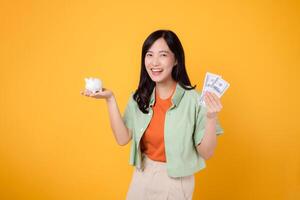 young Asian woman in her 30s, wearing orange shirt and green jumper, revealing dollar currency and piggy bank on yellow background. Financial money concept. photo