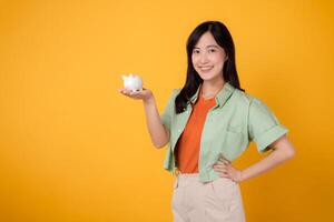 young Asian woman in her 30s, elegantly dressed in orange shirt and green jumper, revealing piggy bank while striking an akimbo gesture on yellow background. Financial money concept. photo