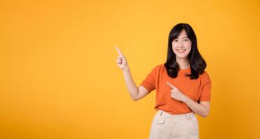 Feel the joy with a young Asian woman in her 30s, donning an orange shirt, pointing to free copy space on yellow background. recommending product concept. photo