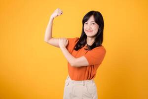 abrazo confianza y empoderamiento como un joven asiático mujer en su 30s levanta un puño arriba mano firmar gesto, vistiendo un naranja camisa en amarillo antecedentes. empoderamiento y feminismo concepto. foto