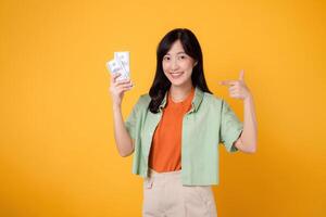 young 30s asian woman happy face dressed in orange shirt and green jumper pointing finger to dollar currency isolated on yellow background. finance business concept. photo