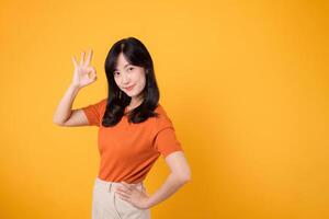 Embody joy with a young Asian woman in her 30s, donning an orange shirt, giving a okay hand gesture on yellow background. product recommending concept. photo