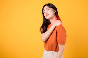 Young asian woman 30s wearing orange shirt holding her pain shoulder isolated on yellow background. office syndrome health care concept. photo