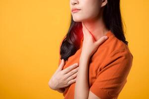 cuidando joven asiático mujer en su 30, vistiendo un naranja camisa, sostiene su dolor cuello en amarillo antecedentes. foto