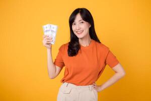 Radiant young Asian woman in her 30s, holding cash money dollars, standing on vibrant yellow backdrop. photo