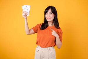 Cheerful Asian woman in her 30s points to cash money dollars on vibrant yellow backdrop. Wealth and success concept. photo