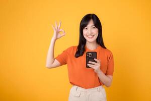 Smiling Asian woman in her 30s, wearing orange shirt, using smartphone with okay hand sign on vibrant yellow background. New mobile app concept. photo