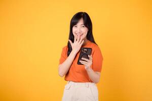 Captivated Asian woman in her 30s, wearing orange shirt, using smartphone with fist up hand sign on vibrant yellow background. Enchanting new app journey. photo