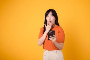 Amazed Asian woman in her 30s, wearing orange shirt, using smartphone with fist up hand sign on vibrant yellow background. Exciting new app discovery. photo