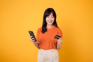 Happy young Asian woman 30s in orange shirt, using smartphone and holding credit card on yellow background. Seamless online shopping. photo