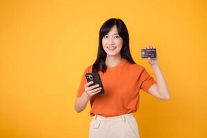 Dynamic young Asian woman in her 30s, using smartphone and holding credit card on yellow background. Secure online payment. photo