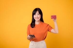 alegre asiático mujer en su 30, utilizando teléfono inteligente y demostración crédito tarjeta en amarillo antecedentes. suave en línea compras. foto