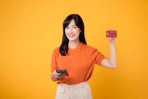 alegre asiático mujer en su 30, utilizando teléfono inteligente y demostración crédito tarjeta en amarillo antecedentes. seguro en línea compras. foto