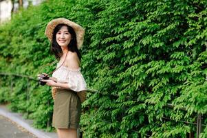 retrato de asiático joven mujer viajero con Costura sombrero y cesta y un cámara en verde público parque naturaleza antecedentes. viaje viaje estilo de vida, mundo viaje explorador o Asia verano turismo concepto. foto