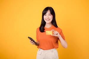 Fresh Asian woman in her 30s, using smartphone and presenting credit card on vibrant yellow background. Speedy online shopping payment transaction. photo