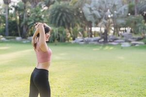 Embrace a wellness healthy lifestyle with back portrait of 30s Asian woman in pink sportswear, breathing fresh air and preparing body before running in public park at sunset. fitness outside concept photo